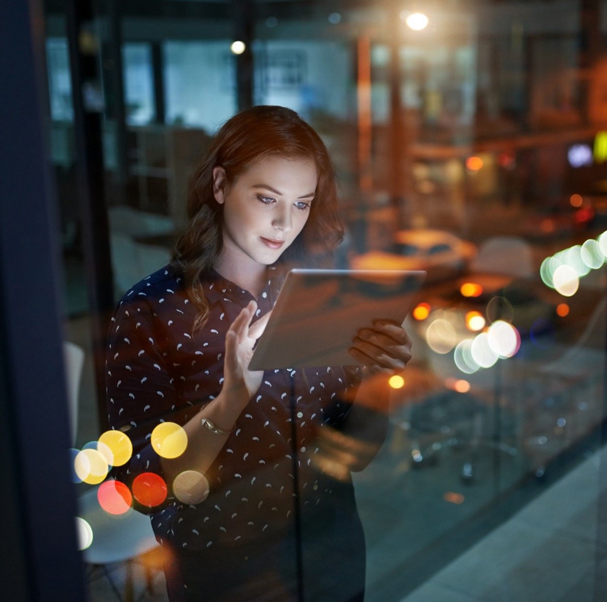 A woman reviewing a tablet at night with a city street visible through the glass behind her.
